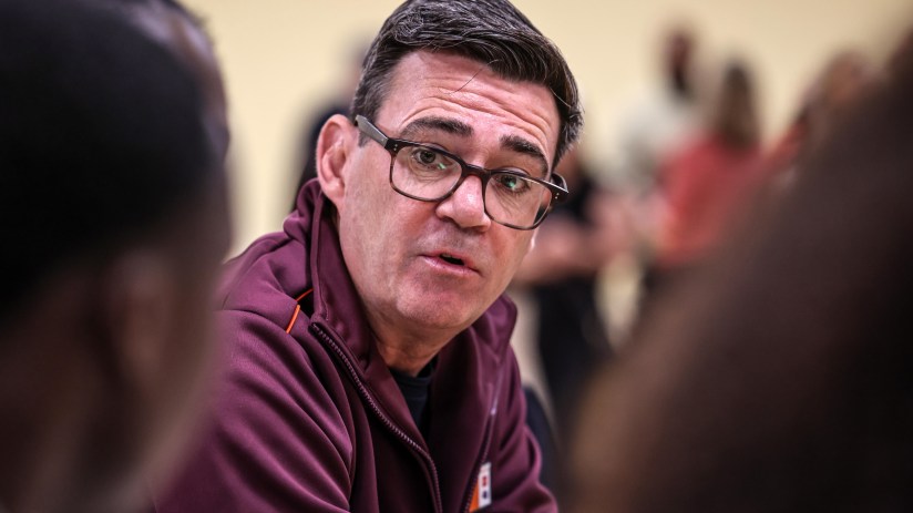 Man in glasses and maroon jacket speaking, with out-of-focus figures in the background.