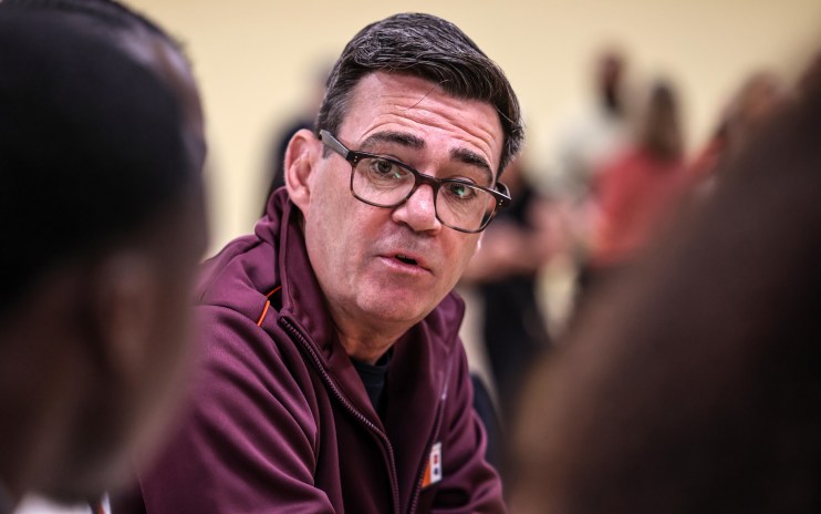Man in glasses and maroon jacket speaking, with out-of-focus figures in the background.
