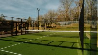 Outdoor padel tennis court with green turf, high fences, and a net, against a backdrop of trees and buildings.