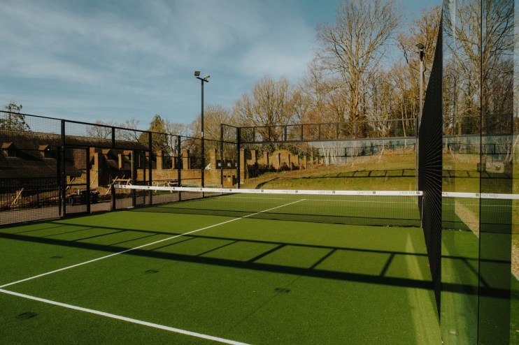 Outdoor padel tennis court with green turf, high fences, and a net, against a backdrop of trees and buildings.