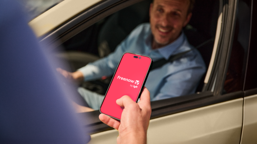 Hand holding a smartphone displaying the FreeNow by Lyft app, with a smiling driver in a car background