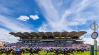 Horses and jockeys racing past a grandstand packed with spectators at the Qatar Goodwood Festival under a blue sky