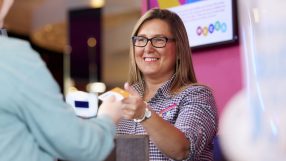 Smiling female receptionist with glasses handing a card to a customer at a bright reception desk.