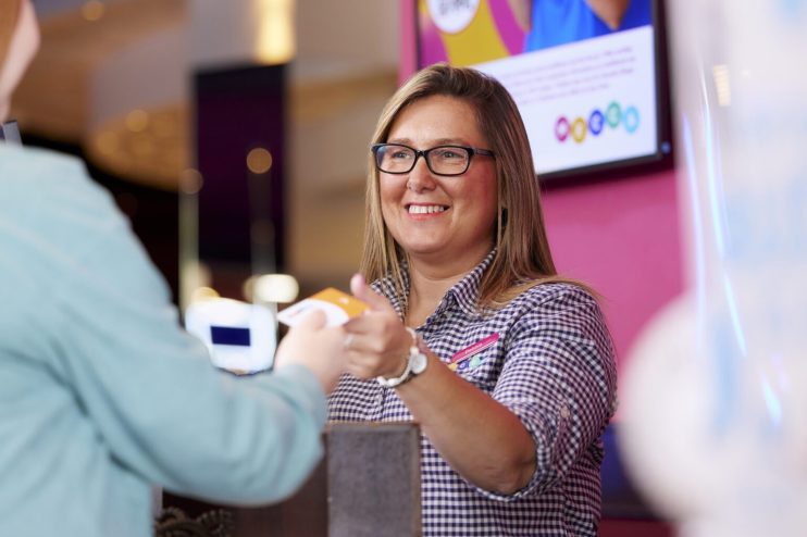 Smiling female receptionist with glasses handing a card to a customer at a bright reception desk.