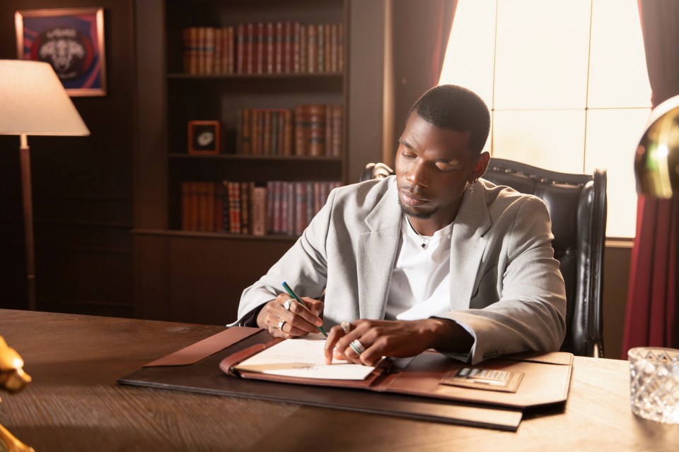 Paul Pogba signing documents at a dark wooden desk in a room with a bookshelf and lamp.