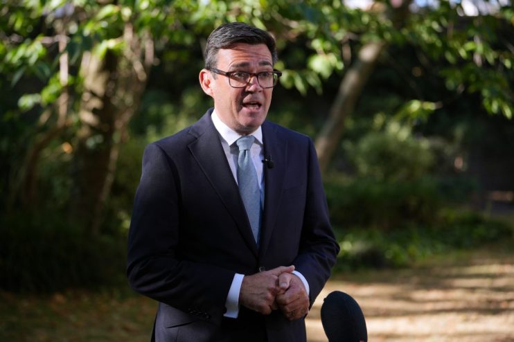 Andy Burnham, wearing a dark suit and glasses, speaks outdoors with trees in the background.