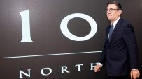 Andy Burnham in a dark suit and glasses, standing before a 10 NORTH graphic display.