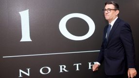Andy Burnham in a dark suit and glasses, standing before a 10 NORTH graphic display.