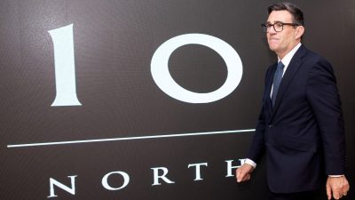 Andy Burnham in a dark suit and glasses, standing before a 10 NORTH graphic display.