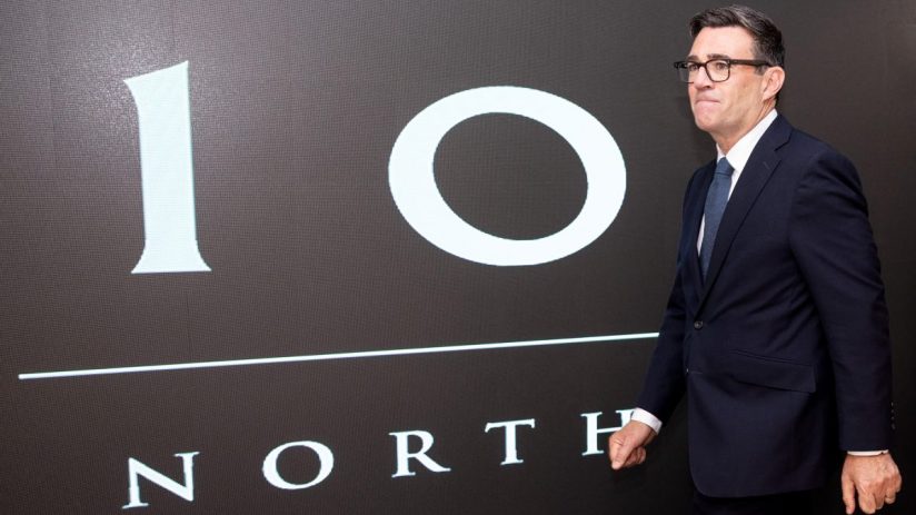 Andy Burnham in a dark suit and glasses, standing before a 10 NORTH graphic display.