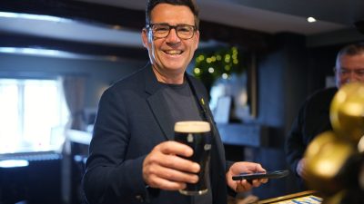 Andy Burnham smiling and holding a pint of beer and a smartphone in a pub setting