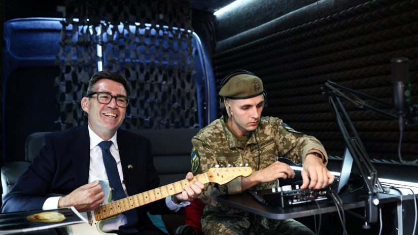 Andy Burnham smiling and playing guitar in Ukraine next to a soldier in uniform adjusting audio equipment