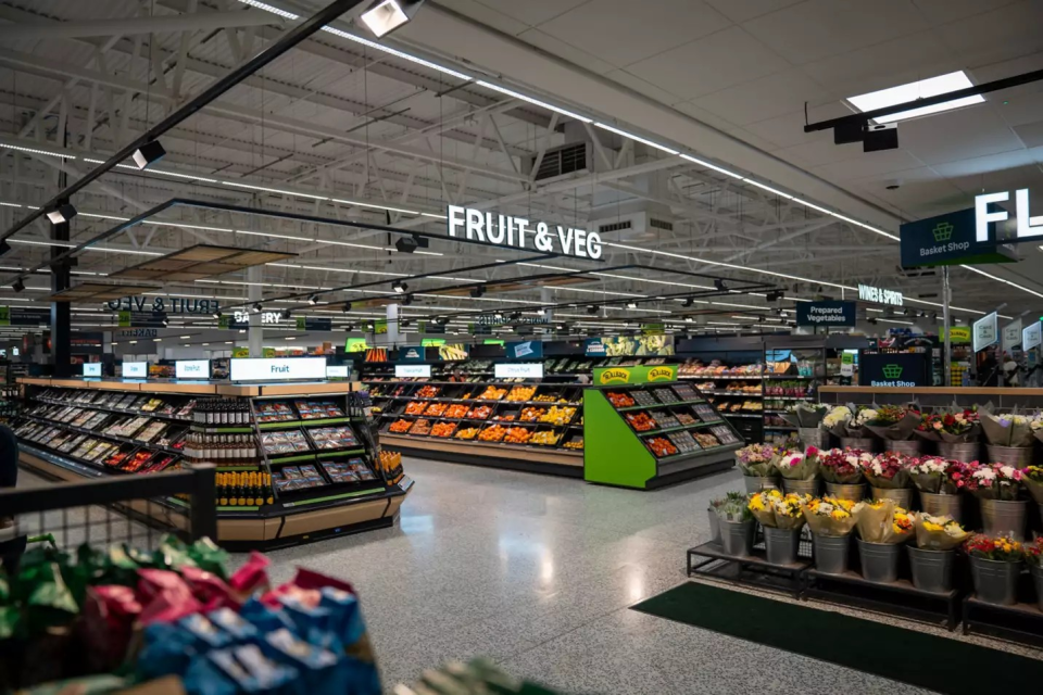 Asda supermarket aisle with Fruit & Veg sign, fresh produce displays, and flowers, reflecting Q2 trading