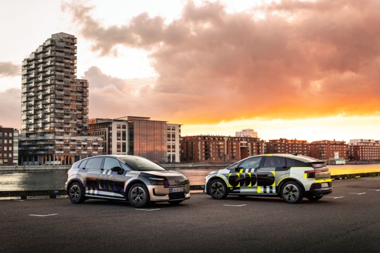 Two Audi A2 concept cars with distinctive designs parked by a waterfront cityscape at sunset.