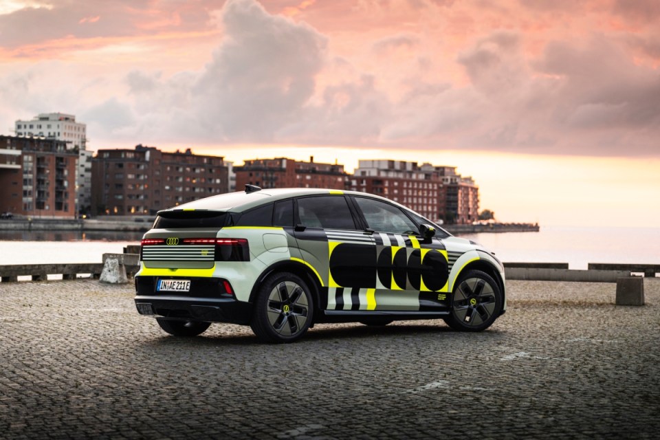 Audi A2 electric concept car with geometric camouflage livery parked on cobblestones at sunset, urban waterfront in backgr...