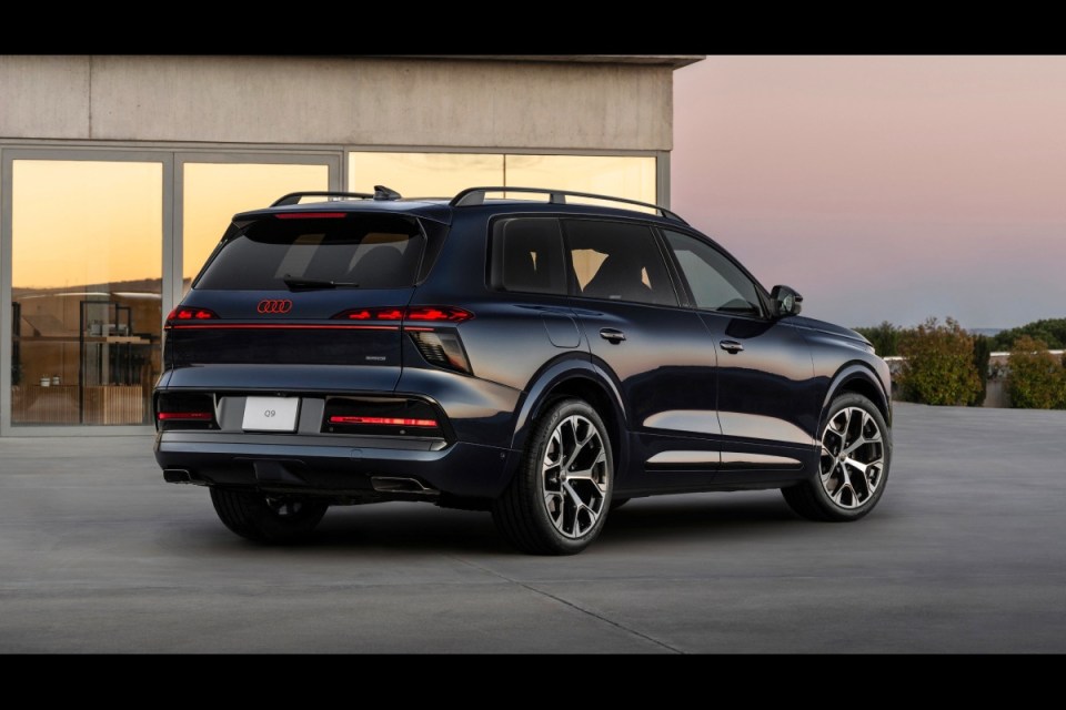 Dark blue Audi Q9 SUV, rear-side view, parked outdoors at dusk with a modern building background.