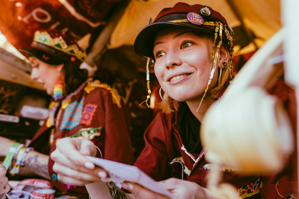 Smiling Boomtown 2026 actor in a red cap with sequins and badges, looking right.
