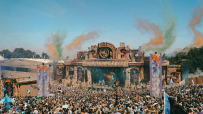 Large crowd at Boomtown festival main stage with vibrant smoke and confetti, during a sunny day.
