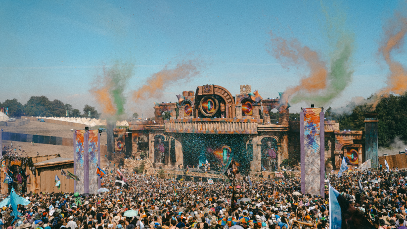 Large crowd at Boomtown festival main stage with vibrant smoke and confetti, during a sunny day.