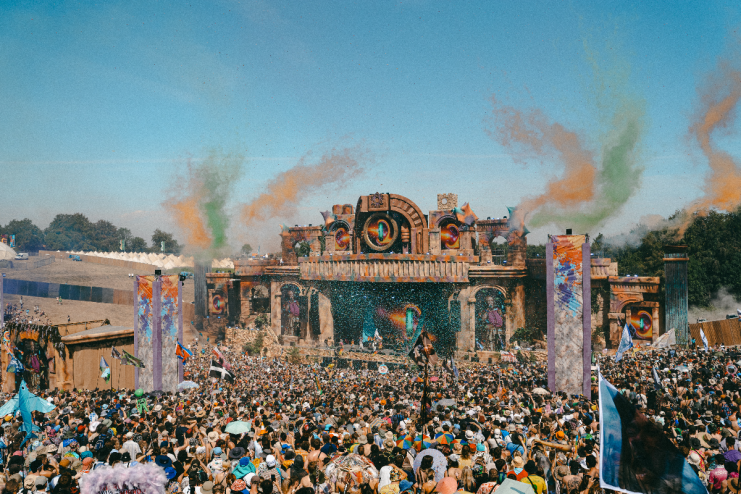 Large crowd at Boomtown festival main stage with vibrant smoke and confetti, during a sunny day.
