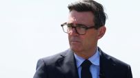 Andy Burnham, Mayor of Greater Manchester, in a suit and glasses, looking serious against a bright sky.