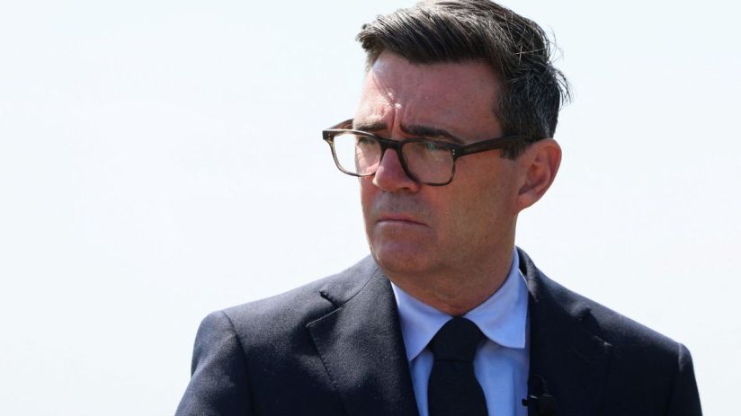 Andy Burnham, Mayor of Greater Manchester, in a suit and glasses, looking serious against a bright sky.