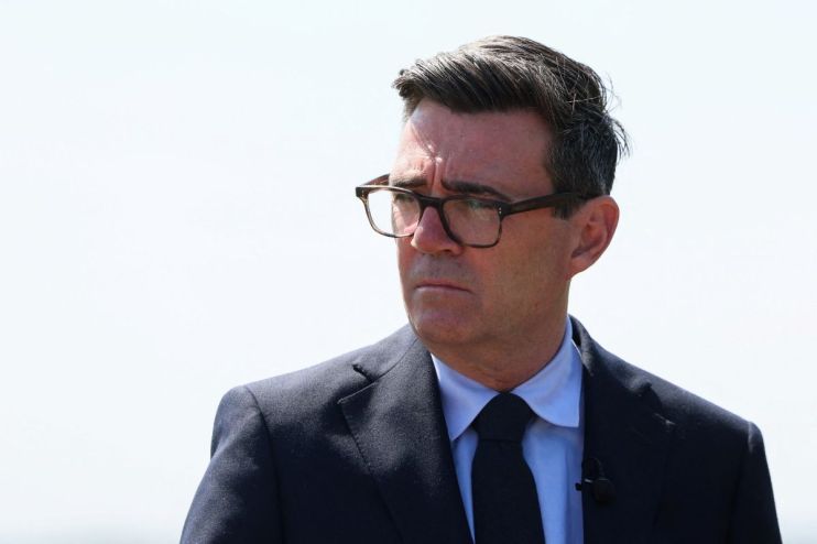 Andy Burnham, Mayor of Greater Manchester, in a suit and glasses, looking serious against a bright sky.