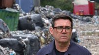 Andy Burnham, Mayor of Greater Manchester, in a dark jacket and glasses, standing before a large pile of waste.