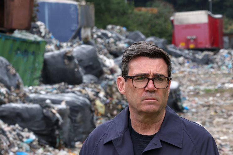 Andy Burnham, Mayor of Greater Manchester, in a dark jacket and glasses, standing before a large pile of waste.