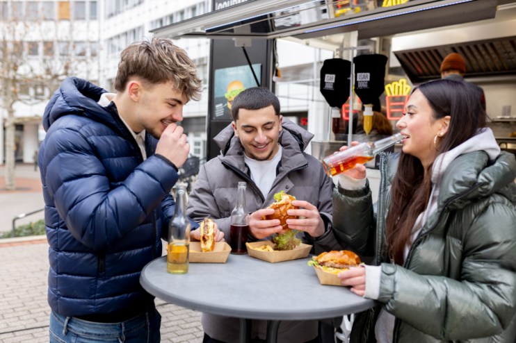 Three young adults enjoying chicken burgers and drinks from a food truck, casually dining outdoors.