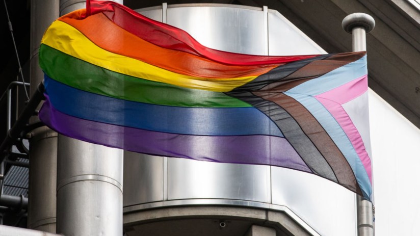 Progress Pride flag flying on a pole against a modern building, symbolizing trans policies in city firms