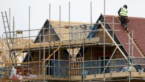 Construction worker in high-visibility vest on a new house roof with red tiles, surrounded by scaffolding.
