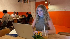 Anna, a young woman, smiles while typing on a laptop at a cafe table with a drink and potted plant.