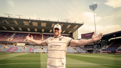 Smiling man in Burnley FC kit with Finotive One sponsor, arms outstretched, on a football pitch with stadium stands behind.