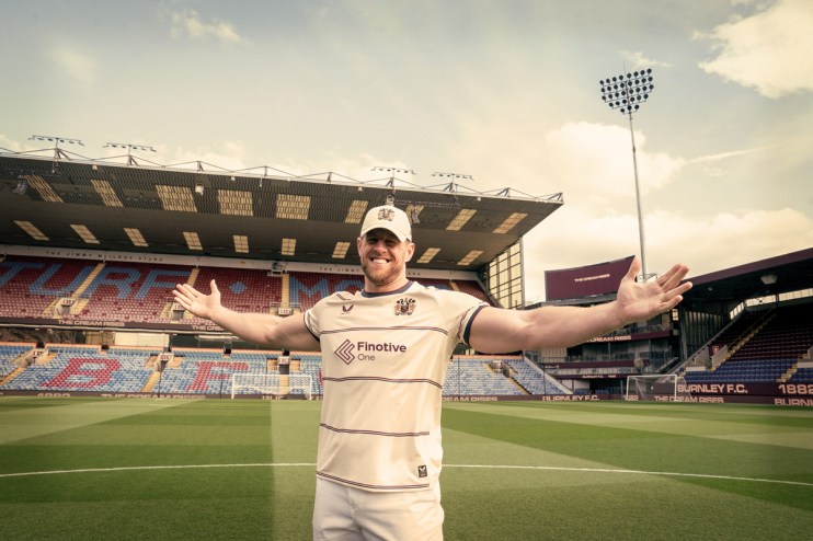 Smiling man in Burnley FC kit with Finotive One sponsor, arms outstretched, on a football pitch with stadium stands behind.
