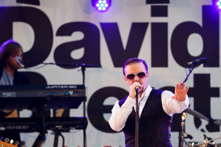 Ricky Gervais as David Brent performing on stage, wearing sunglasses and a patterned vest.