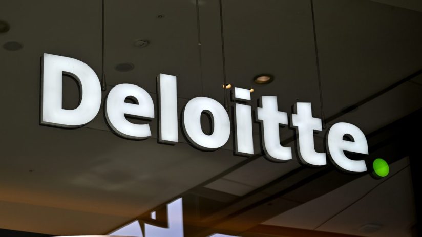 Illuminated white Deloitte logo sign with a green dot, mounted on a dark background, reflecting in a window.