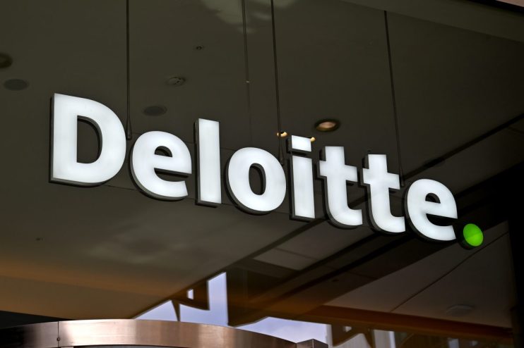 Illuminated white Deloitte logo sign with a green dot, mounted on a dark background, reflecting in a window.