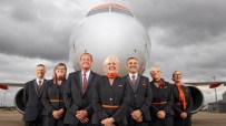 Six smiling EasyJet cabin crew, 50+, in uniform in front of an airplane on a cloudy day.