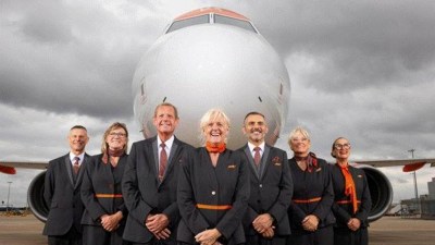 Six smiling EasyJet cabin crew, 50+, in uniform in front of an airplane on a cloudy day.
