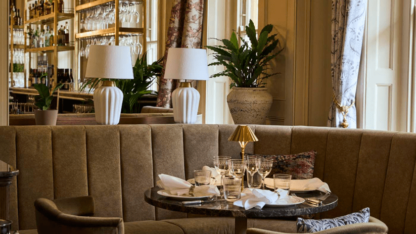 Emberwood Bath restaurant interior with set table, brown banquette seating, white lamps, and a bar in the background.
