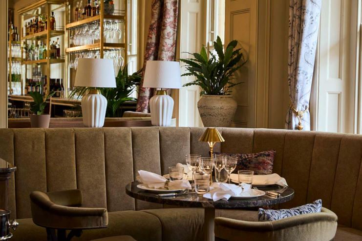 Emberwood Bath restaurant interior with set table, brown banquette seating, white lamps, and a bar in the background.