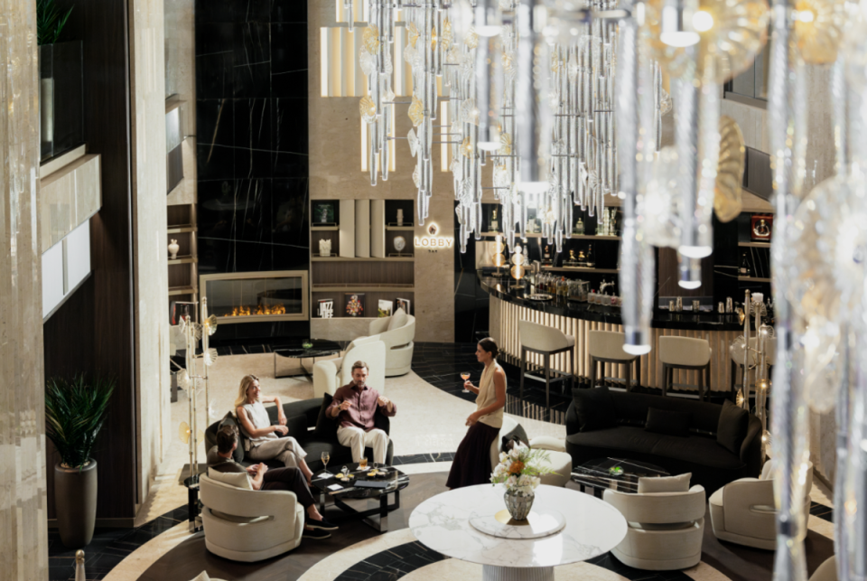 Elegant hotel lobby with guests relaxing, a bar, fireplace, and modern chandelier.