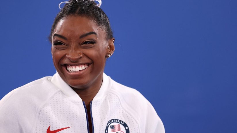 Simone Biles smiling, wearing a white US Olympic Team jacket with the Nike logo, against a blue background.