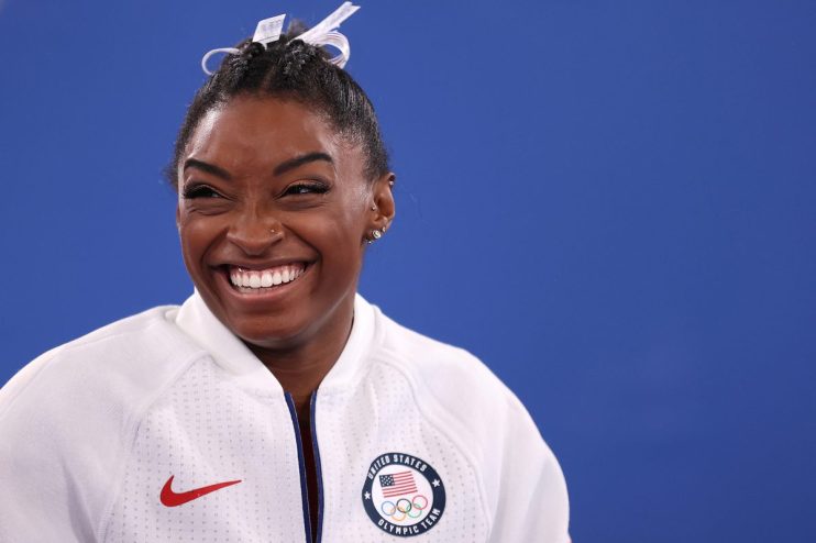 Simone Biles smiling, wearing a white US Olympic Team jacket with the Nike logo, against a blue background.