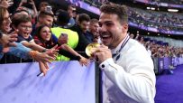 Smiling athlete in white jacket holding a gold medal, reaching out to cheering fans in a stadium.
