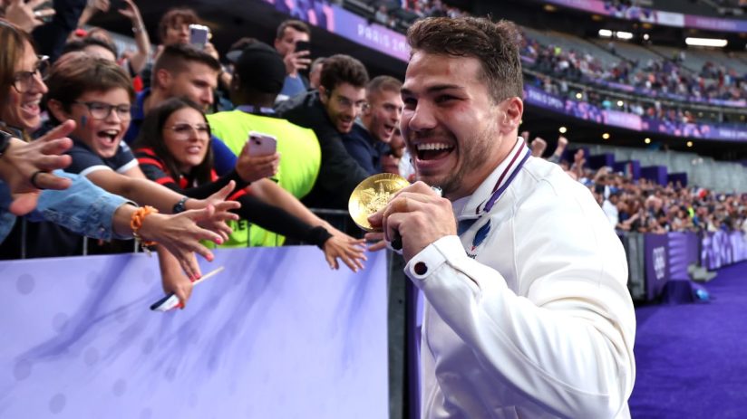 Smiling athlete in white jacket holding a gold medal, reaching out to cheering fans in a stadium.