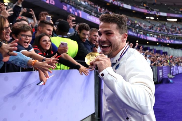 Smiling athlete in white jacket holding a gold medal, reaching out to cheering fans in a stadium.
