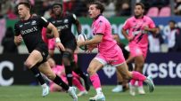 Rugby player in a pink uniform running with the ball, pursued by an opponent in a black jersey.