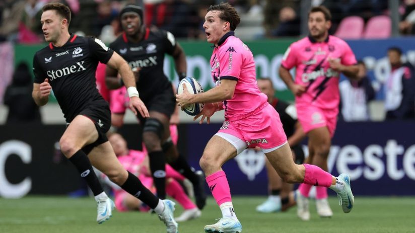 Rugby player in a pink uniform running with the ball, pursued by an opponent in a black jersey.